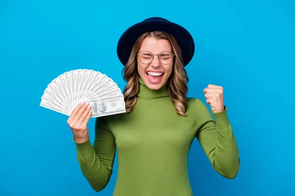 Photo de joyeux fou bouclé dame tenir fan dollar dollars argent personne riche salaire indépendant succès femme d'affaires soulever poing usure spécifications chapeau vert col roulé isolé fond de couleur bleue — Photo