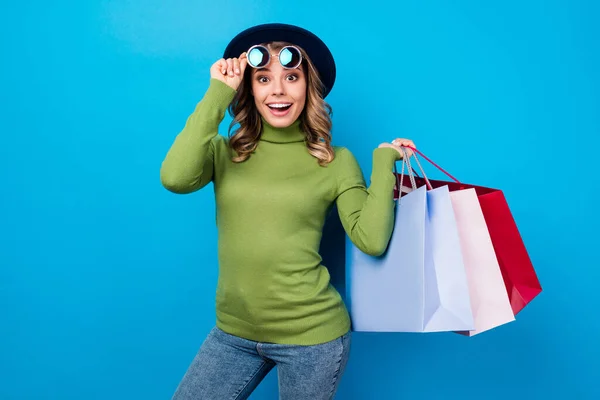 Φωτογραφία του χαρούμενα αστεία σγουρά κυρία κατέχουν πολλές τσάντες κατάστημα πακέτα εθισμένος shopaholic ενθουσιασμένοι διάθεση φορούν ήλιο specs καπέλο casual πράσινο τζιν ζιβάγκο απομονωμένο μπλε χρώμα φόντο — Φωτογραφία Αρχείου