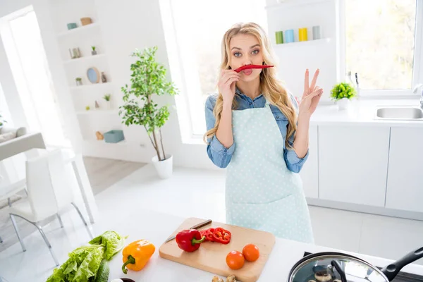 Foto van vrolijke huisvrouw blond dame gesneden salade koken smakelijk diner ontbijt houden rode hete chili peper als snor tonen v-teken symbool speels goed humeur staan moderne keuken binnen — Stockfoto
