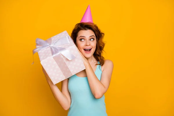 Foto van vrij grappige dame houden grote giftbox handen onverwachte cadeau proberen te raden wat binnen dragen roze papieren verjaardagskap casual teal tank-top geïsoleerde heldere gele kleur achtergrond — Stockfoto