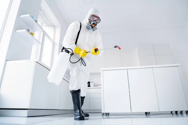 Low angle view full body foto van gefocuste schonere man in beschermende uniform bril handschoenen houden sprayer spray stop covid19 epidemie verspreiden oppervlakte vloer in keuken huis binnen — Stockfoto