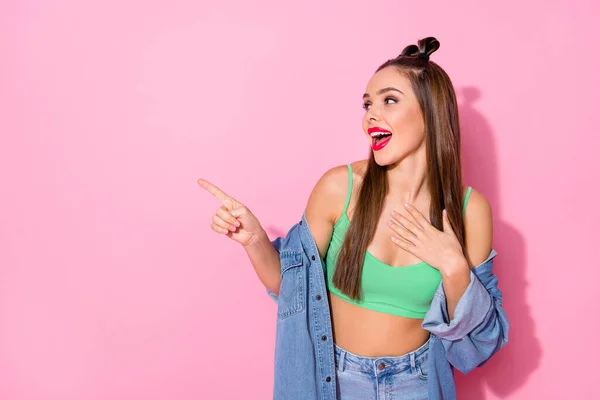 Photo of pretty funky lady red lips positive emotions directing finger side empty space novelty wear denim jacket green crop top naked belly isolated pastel pink color background