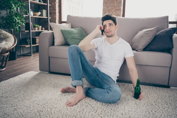 Retrato de su agradable chico deprimido triste atractivo sentado en la alfombra hablando por teléfono con un amigo bebiendo cerveza en el moderno loft industrial de ladrillo de estilo interior sala de estar en interiores — Foto de Stock