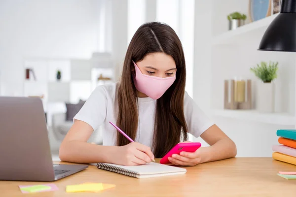 Foto van kleine mooie leerling kleine school dame laptop notebook online les zit bureau gebruik maken van veiligheid gezichtsmasker schrijven essay zoeken telefoon website afstand quarantaine studie woonkamer binnen — Stockfoto