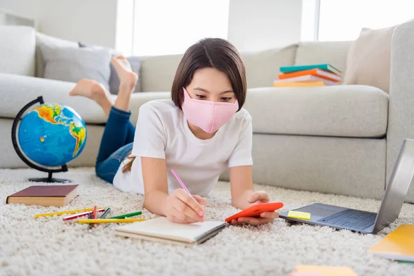 Foto van een kleine leerling school meisje zoeken telefoon informatie voor te bereiden notebook online les leraar liggen vloer binnenlandse sociale afstand quarantaine studie ademhalingsmasker binnenshuis — Stockfoto