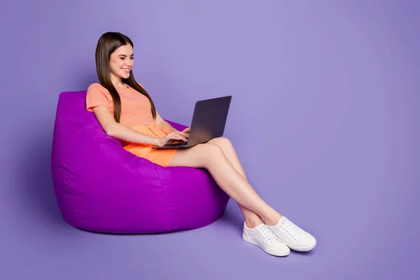 Foto profil lengkap dari wanita ceria duduk nyaman beanbag browsing notebook pekerjaan freelance mengenakan bergaris-garis oranye rok mini T-shirt terisolasi pastel warna ungu latar belakang — Stok Foto