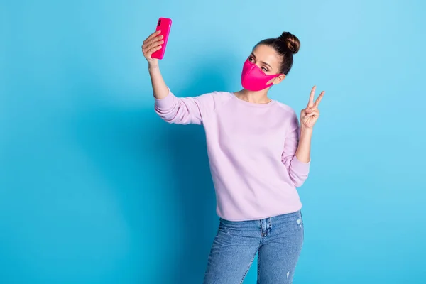 Foto de senhora muito atraente manter distância social pessoas segurar telefone fazendo selfies on-line blog mostrar v-sinal símbolo desgaste proteger rosto máscara jeans pulôver isolado azul cor de fundo — Fotografia de Stock