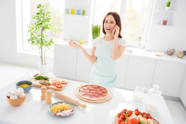 Foto van mooie vrolijke huisvrouw genieten hobby koken familie recept diner pizza spreken telefoon quarantaine tijd thuis blijven dragen t-shirt schort staande moderne keuken binnen — Stockfoto