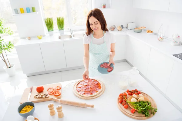 Foto van mooie vrolijke huisvrouw genieten hobby bereiden familie recept zet salami worstjes plakjes op rij deeg Italiaanse pizza quarantaine tijd thuis blijven moderne keuken binnen — Stockfoto