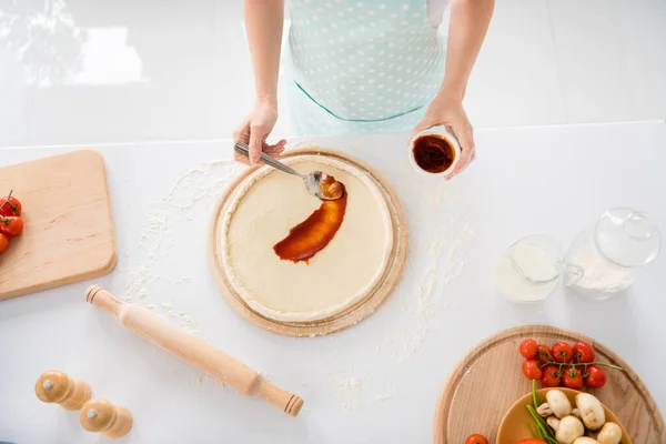 Top above high angle view cropped photo of housewife woman make pizza spread tomato sauce seasoning on dough wear dotted apron in kitchen house indoors