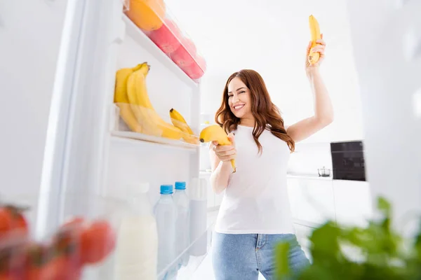Фото красивої домогосподарки, яка відкриває двері холодильника Їжа перекушування дієти ранкових вихідних, тримають два банани, які стріляють, як зброя яскрава світла кухня в приміщенні повсякденний одяг — стокове фото