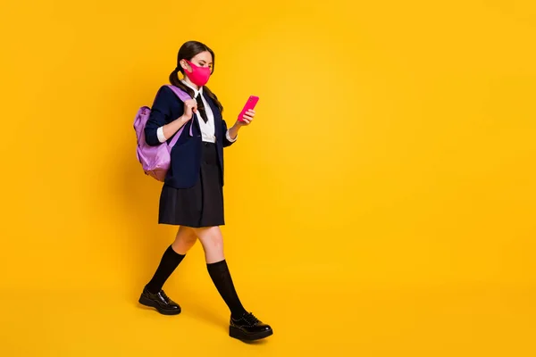 Full size profil boczny zdjęcie liceum nastolatek go chodzić lekcji używać smartfona czat nosić czarny marynarka spódnica długie skarpetki plecak medyczny maska izolowane żółty kolor tło — Zdjęcie stockowe