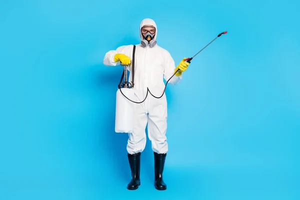 Full length photo medical worker man use equipment bottle disinfect home surface covid19 pathogen infection wear hazmat uniform breathing mask gloves goggles isolated blue color background