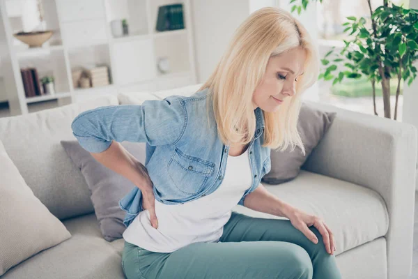 Closeup profile photo of unwell displeased blond mature lady sit couch stay home quarantine hold hand on spine suffer pms touch back massaging injured pain place living room indoors — Stock Photo, Image