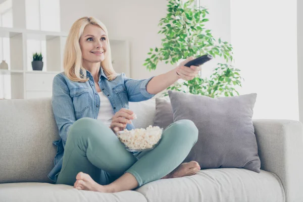 Fotografie atraktivní starší dospělé domácí dáma sedí pohodlné pohovka nohy kříž jíst popcorn dobrou náladu relaxační hodinky tv pobyt doma karanténa čas víkend obývací pokoj uvnitř — Stock fotografie