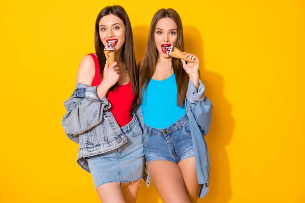 Ritratto di positivo allegro sorelle ragazze divertirsi mangiare leccare delizioso gelato indossare jeans singoletto isolato su sfondo di colore brillante — Foto Stock
