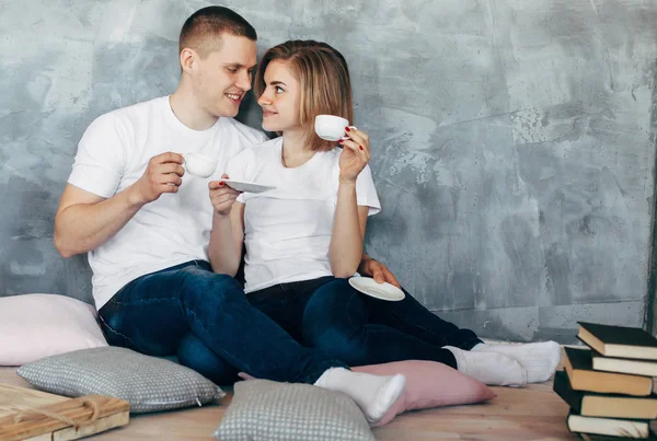 Young loving couple in white t-shirts  sitting on the  floor and  drinking a tea or coffee. Copy space. Grey Background