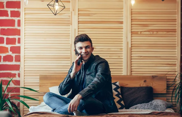 Genç gülümseyen in kapalı atış yatağa oturmuş ve cep telefonuyla konuşuyor dostum. Mutlu genç adam cevap telefon görüşmesi. Kopya alanı