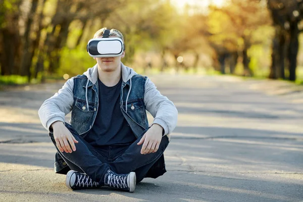 The young blonde man sitting on the road with virtual reality glasses. Outdoor. Copy space.
