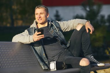 Gerçeklik bulanık satır. Genç sarışın erkek oturur bankta elleri, bir smartphone ile gülümseyen ve göz kulak. Uzay kopyalayın. Açık.