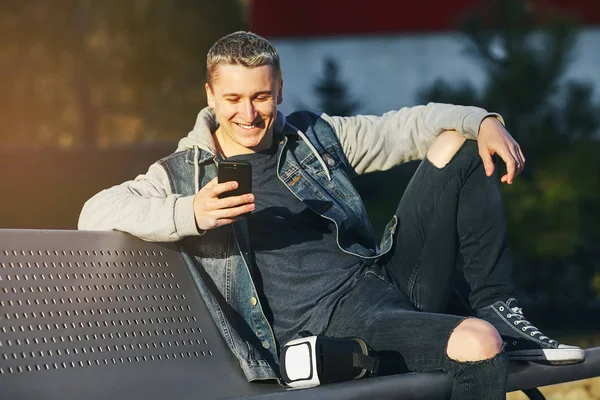 Gerçeklik bulanık satır. Genç sarışın erkek onun smartphone ellerinde tutan bankta oturur ve gülümseme ile iyi eğlenceler. Uzay kopyalayın. Açık.