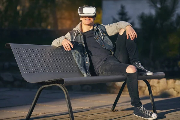 Blurred lines of reality.  The young men sits on the bench and wears VR glasses and looking out and smiling. Outdoor. Copy space.