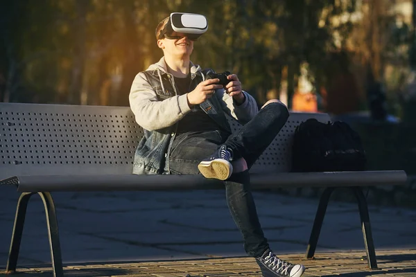 Gerçeklik bulanık satır. Genç erkekler bankta oturur ve Vr gözlük takar ve bir denetleyici elinde tutar ve bir oyun oynuyor. Açık. Kopya alanı.