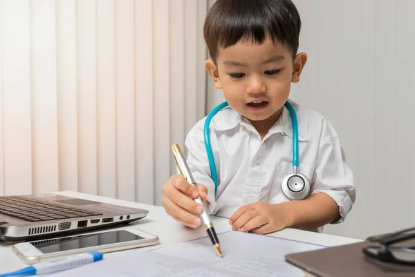 Niño pequeño en uniforme médico usando un bolígrafo en el escritorio —  Fotos de Stock