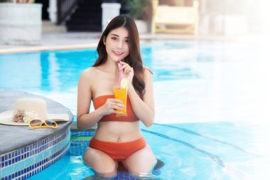 Yazın mayolu seksi Asyalı kadın. Turuncu bikinili güzel bir kadın tropikal bir içkiyle havuzda oturuyor..