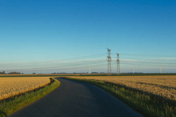 High voltage power lines and transmission towers. Asphalt road passing through agricultural fields in Normandy, France. Electric power industry, road network and agriculture concept