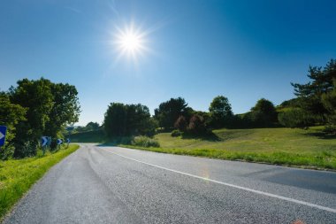 Yeşil alanları ve orman geçerken boş asfalt köy yolunda. Fransa mavi gökyüzünde güneş ışınları ile güneşli kırsal manzara. Taşıma, endüstriyel tarım, yol ağ konsepti.