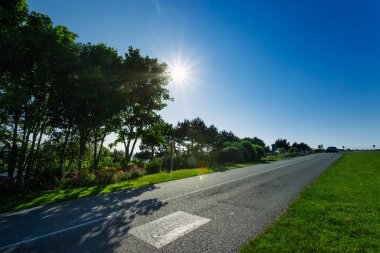 Yeşil alanları ve ormanları geçerken boş asfalt köy yolunda. Kırsal manzara Fransa güneşli bahar gününde. Güneş gökyüzünde. Taşıma, endüstriyel tarım, yol ağ konsepti