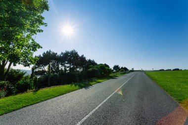 Yeşil alanları ve ormanları geçerken boş asfalt köy yolunda. Kırsal manzara Fransa güneşli bahar gününde. Güneş gökyüzünde. Taşıma, endüstriyel tarım, yol ağ konsepti