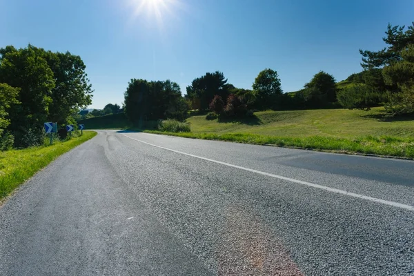 Prázdná země asfaltkou procházející zelené louky a les. Venkovské krajiny na slunečný den s paprsky dopadají na modré obloze ve Francii. Doprava, průmyslové zemědělství, koncepce silniční sítě. — Stock fotografie