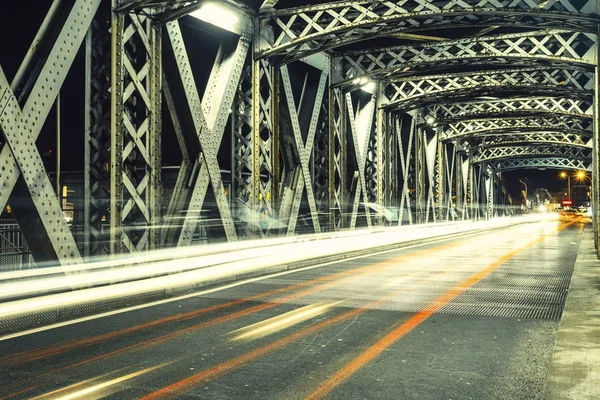 Strada asfaltata sotto la costruzione in acciaio di un ponte in città. Scena urbana notturna con tracce luminose di auto nel tunnel. Tonica — Foto Stock