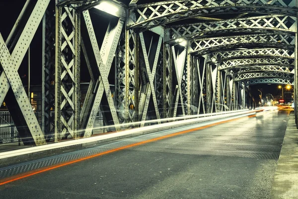 Ασφαλτοστρωμένος δρόμος κάτω από την κατασκευή του χάλυβα από μια γέφυρα στην πόλη. Αστικό τοπίο νύχτα με αυτοκίνητο φως μονοπάτια στη σήραγγα. Ήπια — Φωτογραφία Αρχείου