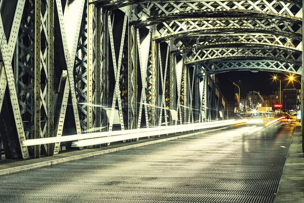 Асфальтовая дорога под стальной конструкцией моста в городе. Ночная городская сцена с автомобильными световыми тропами в туннеле. Toned — стоковое фото