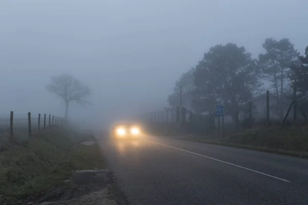 Kraju asfaltu drogi przechodzącej przez las i pola w regionie Normandia, Francja. Wiejski krajobraz w mglisty dzień. Reflektory samochodowe w nocy. Stonowanych — Zdjęcie stockowe