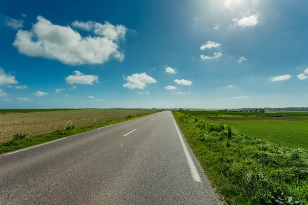 Prázdná země asfaltkou procházející zelené zemědělských polí za slunného dne v regionu Normandie, Francie. Venkovské krajiny s paprsky dopadají na modré obloze, silniční síť a dopravní koncepce — Stock fotografie