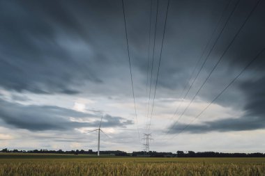 Yüksek gerilim enerji hatları, enerji nakil direkleri ve rüzgar türbinleri bulutlu bir günde tarım alanlarında. Polonyalılar Havai hatları ve siluetleri dusk içinde. Elektrik powere Sanayi ve doğa kavramı.