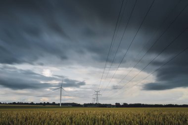 Yüksek gerilim enerji hatları, enerji nakil direkleri ve rüzgar türbinleri bulutlu bir günde tarım alanlarında. Polonyalılar Havai hatları ve siluetleri dusk içinde. Elektrik powere Sanayi ve doğa kavramı.