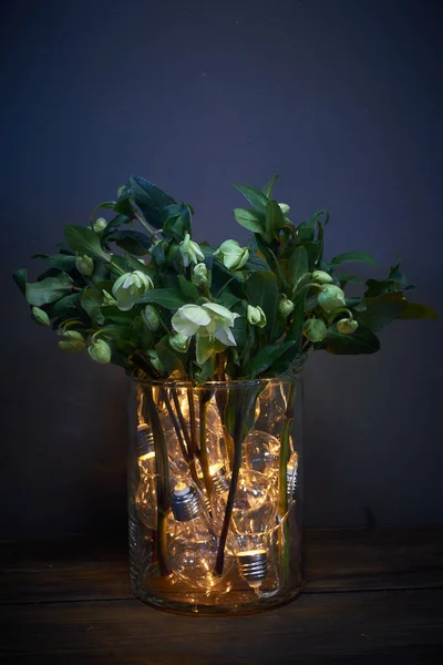 Vaso de vidro cheio de lâmpadas luminosas e um buquê de heléboro, conceito festivo — Fotografia de Stock