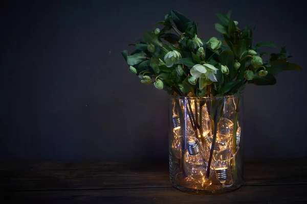 Jarrón de vidrio lleno de bombillas luminosas y un ramo de hellebore, concepto festivo — Foto de Stock
