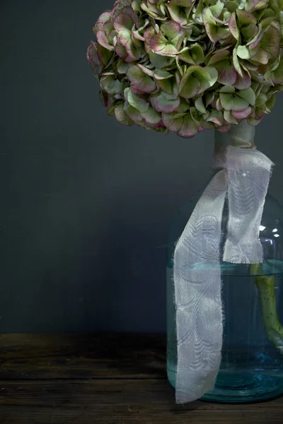 Photo of a huge hydrangea in a glass bottle with a beige ribbon on a dark background, selective focus — Stock Photo, Image