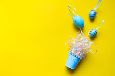 Süslü renkli yumurtalar ve minyatür bir kovada, parlak sarı bir arka planda, minimalist paskalya arkaplanı ya da konsept üzerinde bir kamış yuvası.