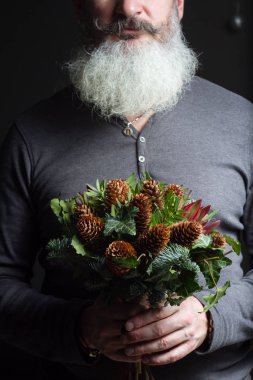 Orta yaşlı sakallı bir adam elinde nobil köknar buketi, kozalak, şam fıstığı, lökodon ve sarmaşık, kış hediyesi konsepti.
