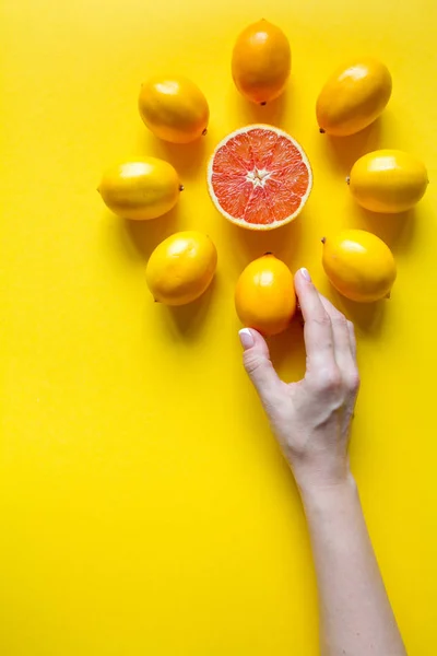 Top view female hand, whole and sliced ripe lemons, grapefruit laid out in the shape of a dial on a yellow surface, concept of health and vitamins