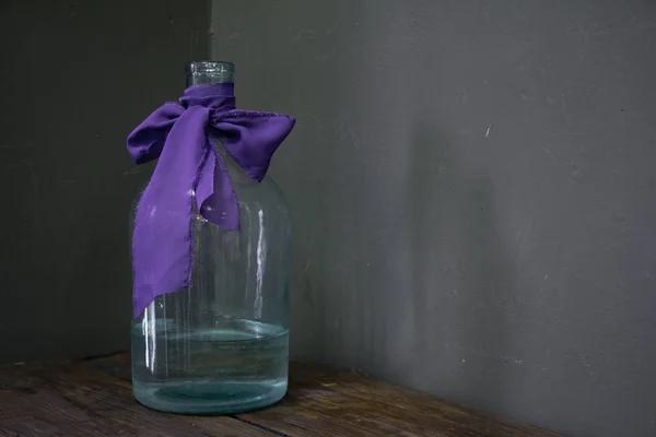 Glass bottle or vase decorated with a purple ribbon on a dark background, selective focus — 스톡 사진