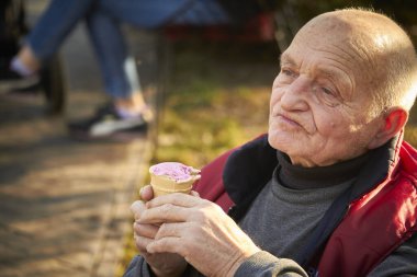 80 yaşında bir adam halka açık bir parkta yürüyor, waffle bardağında dondurma yiyor.