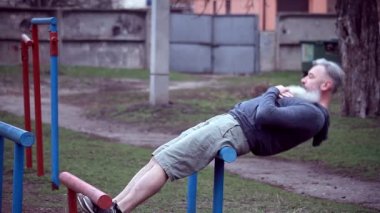 Güçlü kaslı kolları olan yaşlı sakallı bir adam dışarıda egzersiz yapıyor, barfiks çekiyor, yaşlı bir erkek yatay çubukta egzersiz yapıyor.